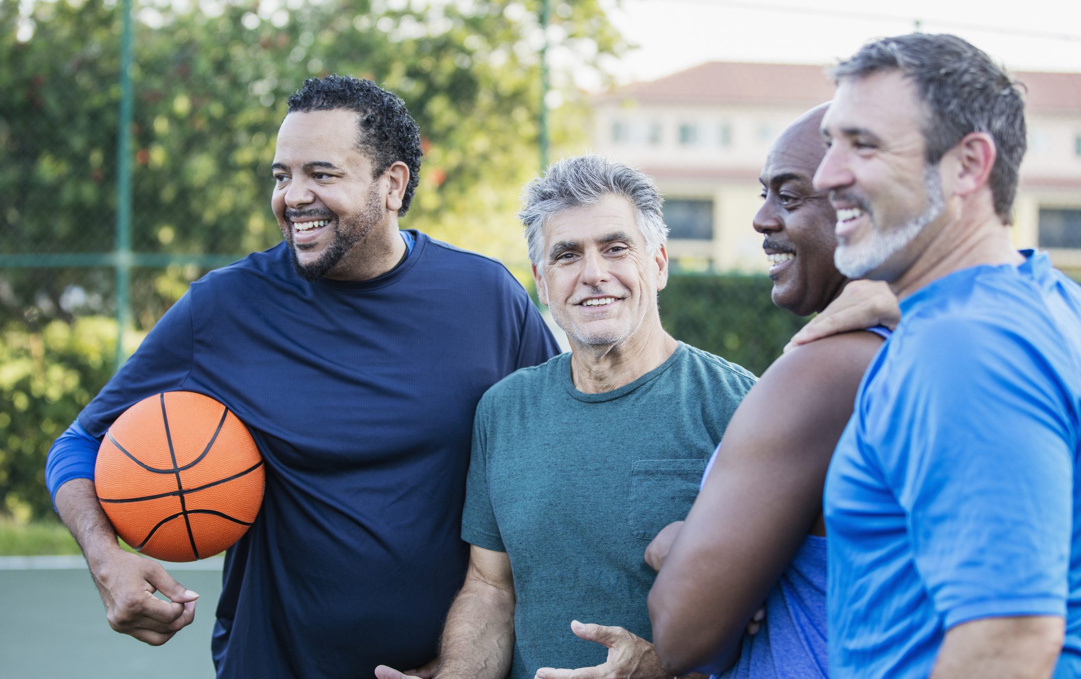 A multi-ethnic group of middle-aged and senior men on an outdoor basketball court. The Caucasian man wearing the green shirt, looking at the camera, is a senior, in his 60s. The bald African-American man is in his 50s, and their friends are mature men in their 40s.