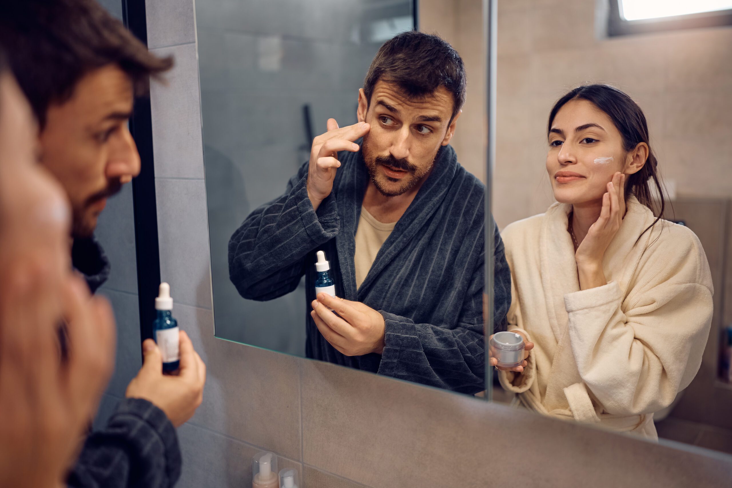Smiling woman and her boyfriend taking care of their facial skin in front of a mirror.